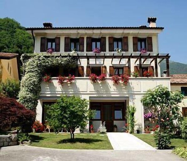 A two-story building with a balcony, a garden in front, and a mountain in the background. - Ernestina (Miane)