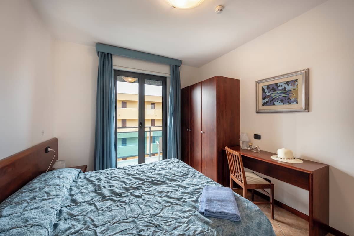 Une chambre d’hôtel comprenant un lit, un bureau, une chaise, une armoire et un balcon avec des rideaux.