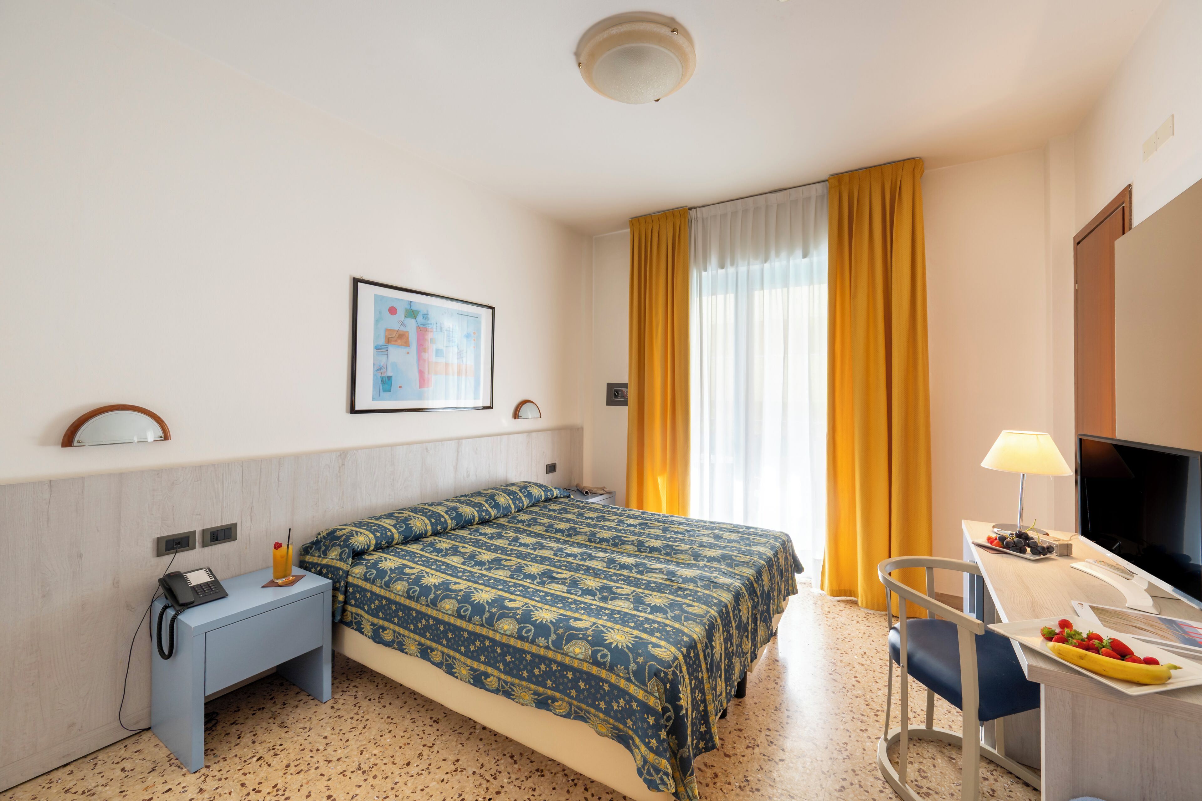 Habitación de hotel con cama, escritorio con computadora, televisor y una pequeña mesa con fruta.