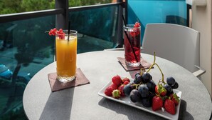 Ein Tisch mit einem Orangensaftglas, einem roten Getränk und einer Schale Erdbeeren und Blaubeeren auf einem Balkon mit Glasgeländer und Blick ins Grüne.