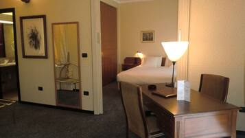 A hotel room with a bed, a desk with a lamp, a chair, a mirror, and a picture on the wall.
