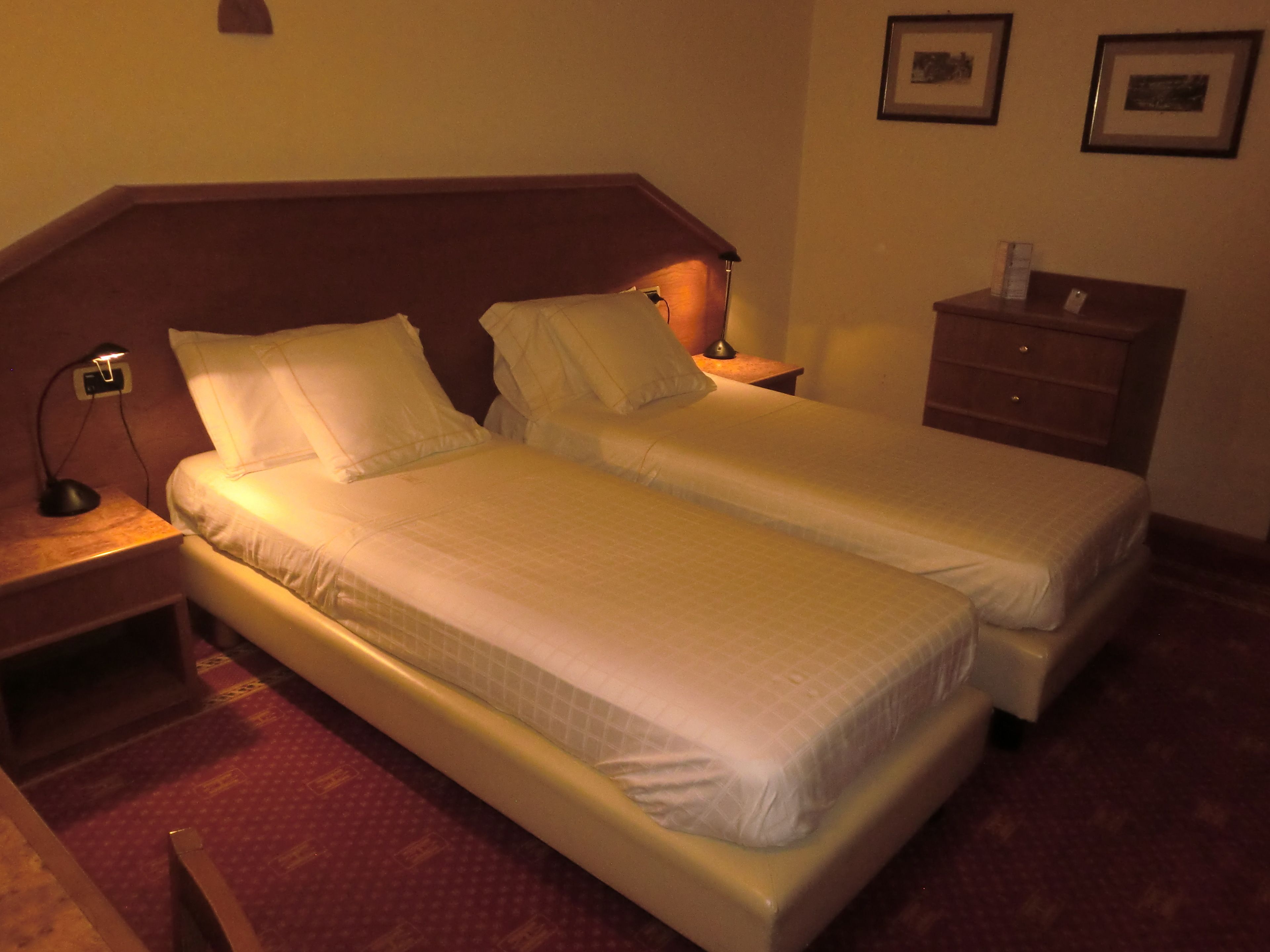 Ein Hotelzimmer mit zwei Einzelbetten, einem Nachttisch mit Lampe, einem Schminktisch und gerahmten Bildern an der Wand.