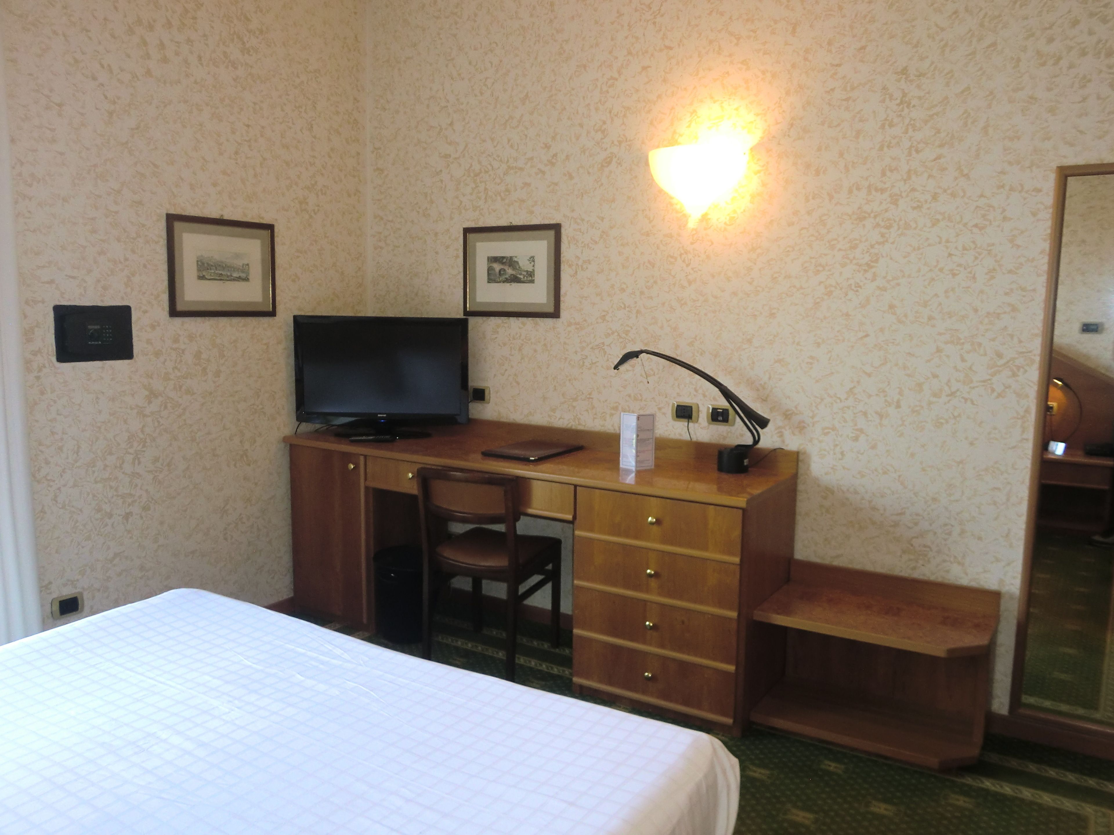 Ein Hotelzimmer mit Bett, Schreibtisch mit Computer, Stuhl, Schreibtischlampe und zwei gerahmten Bildern an der Wand.