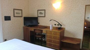 Ein Hotelzimmer mit Bett, Schreibtisch mit Computer, Stuhl, Schreibtischlampe und zwei gerahmten Bildern an der Wand.
