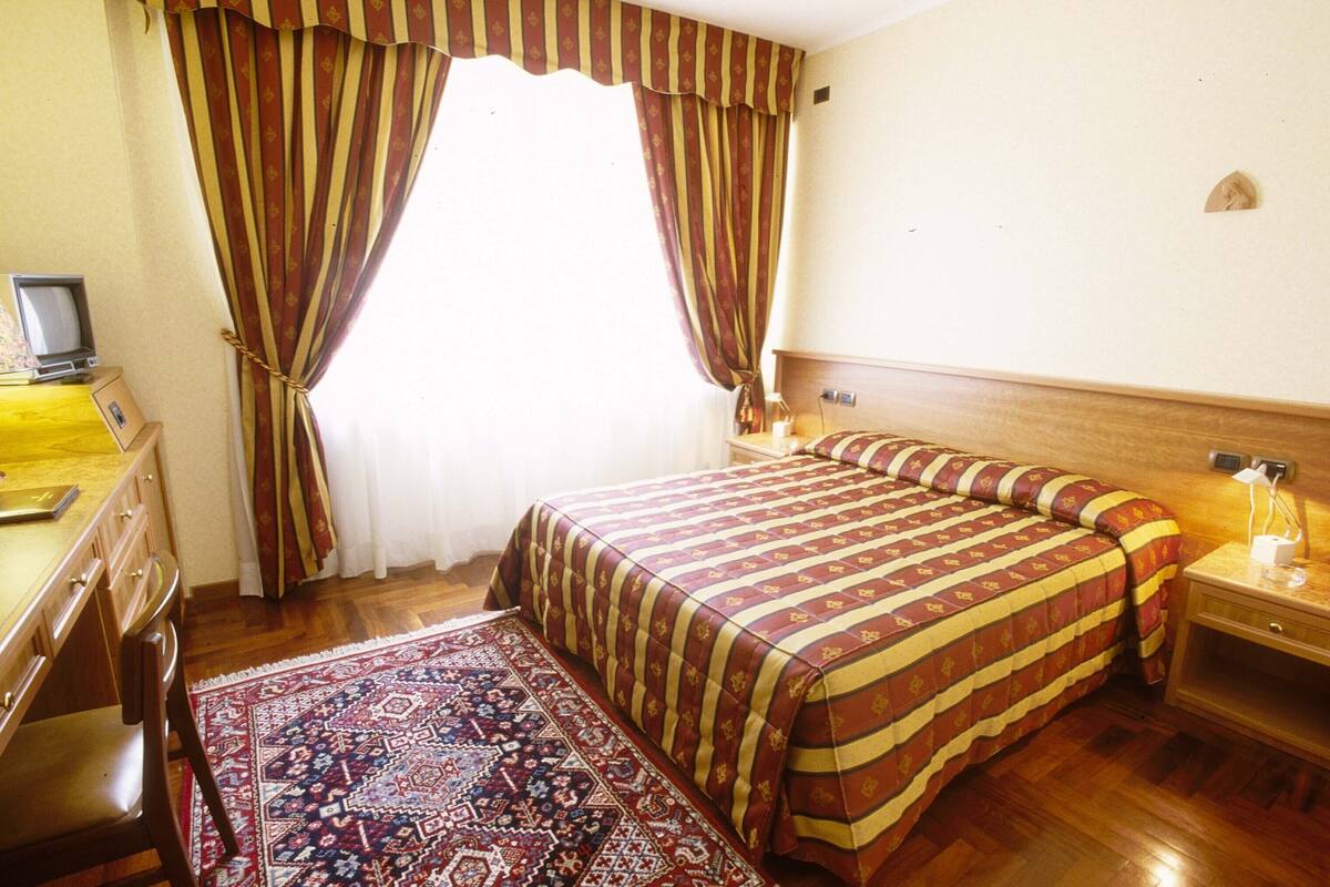 A hotel room with a bed, a desk with a computer, a nightstand with a lamp, and a patterned rug.