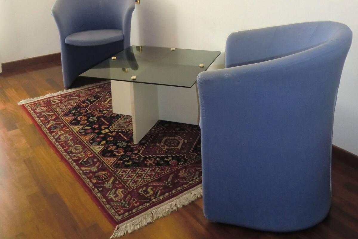 A room with a glass-top table, two blue armchairs, a patterned rug, a framed painting, and a wall-mounted light fixture.
