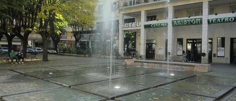 Una plaza pública con una fuente, rodeada de árboles y edificios con letreros de un hotel, cine y teatro.