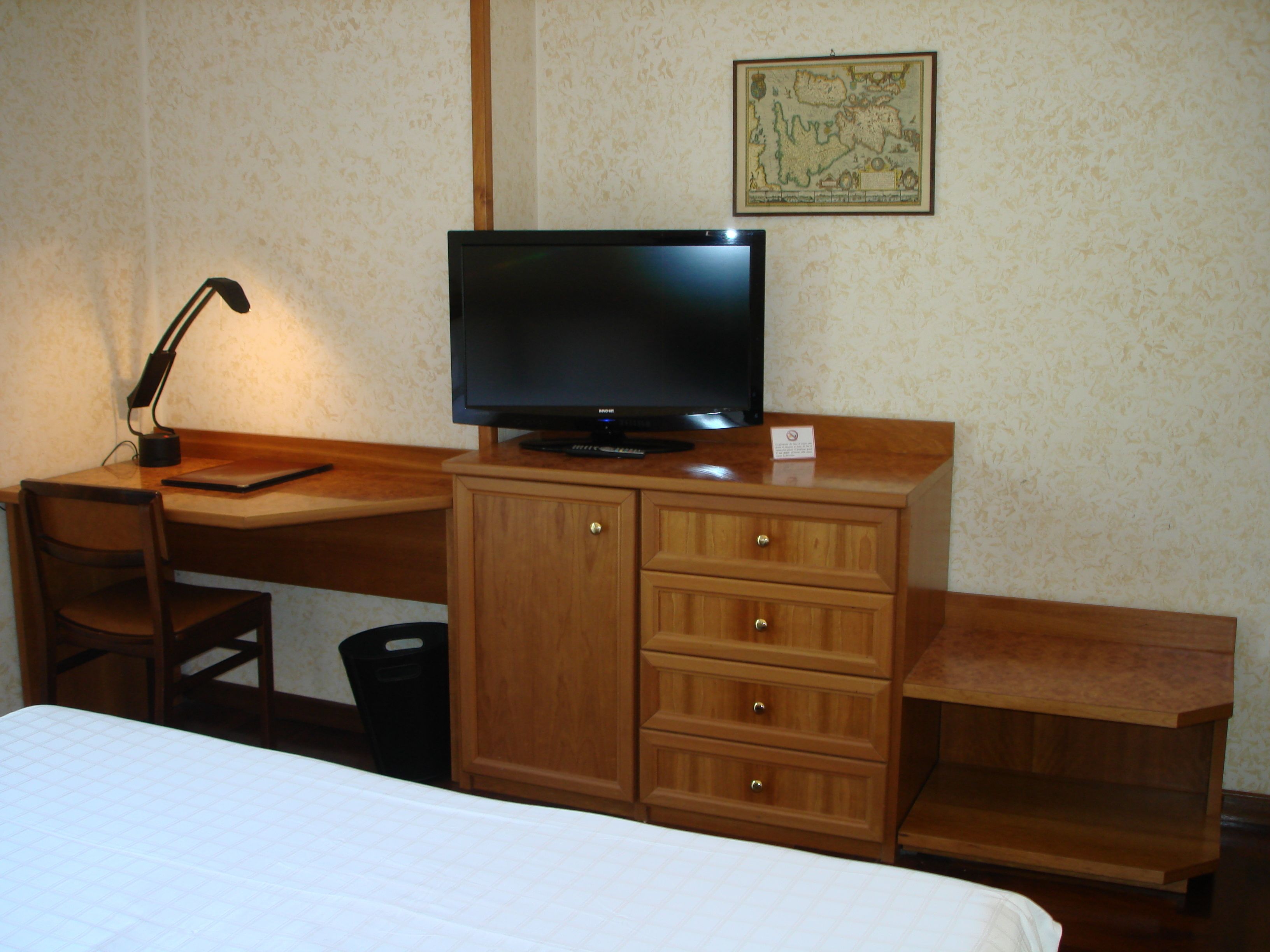 A hotel room with a wooden desk, a television, a chair, a bed, and a map on the wall.