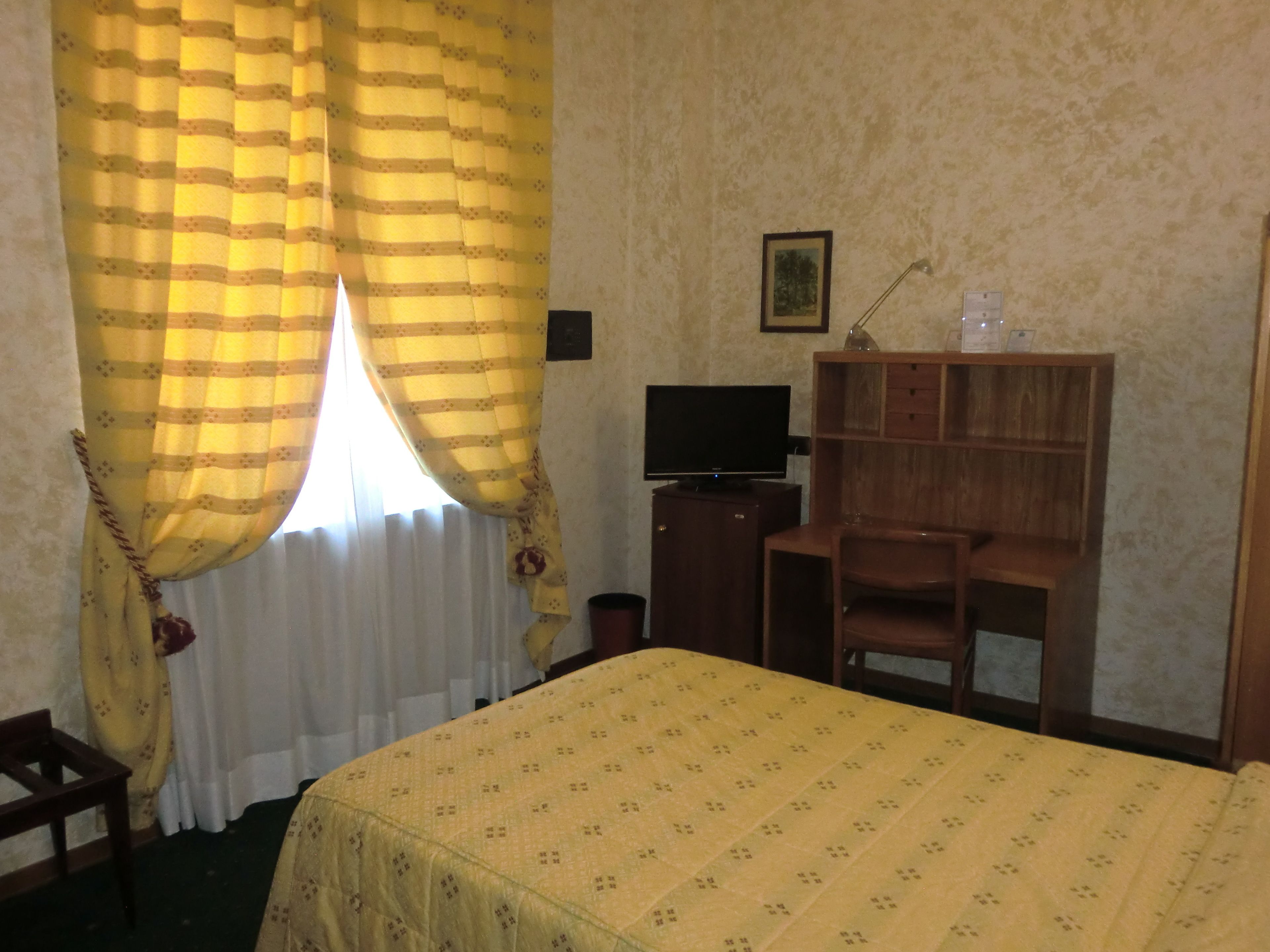 A hotel room with a bed, a television on a wooden cabinet, a desk with a chair, and a window with yellow curtains.