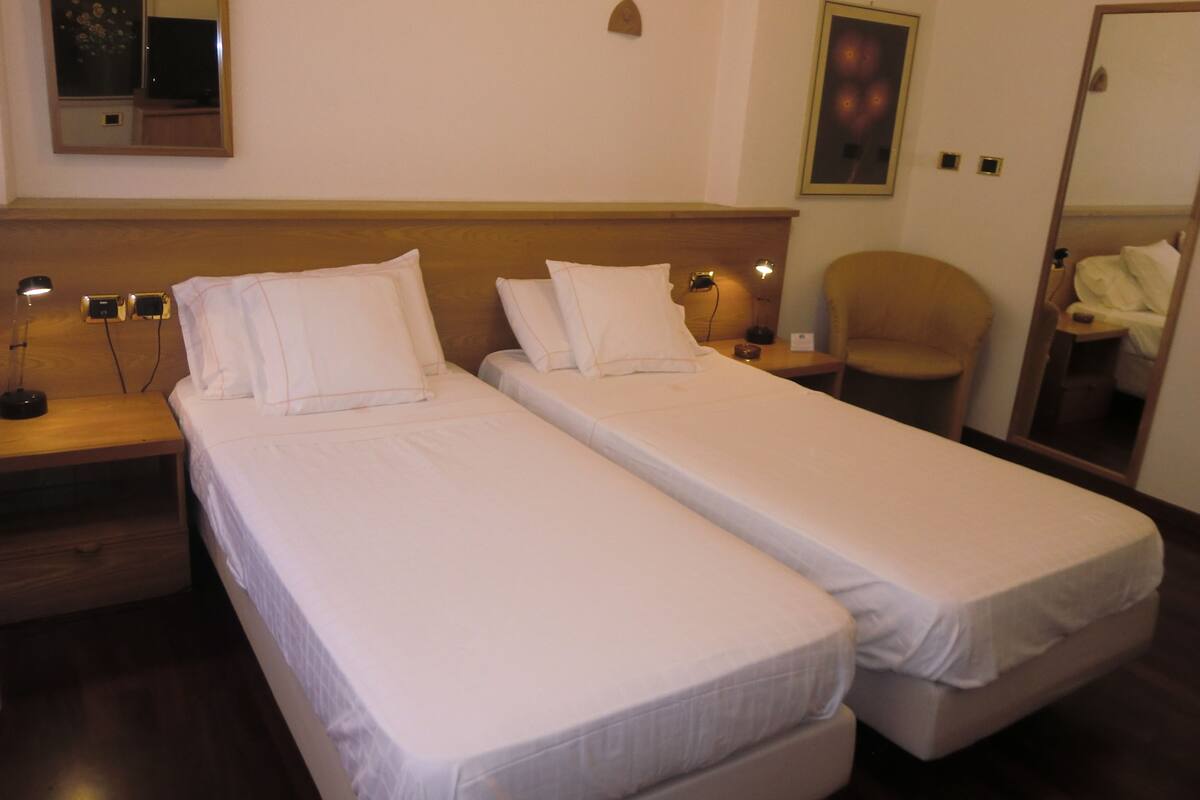 A hotel room with two single beds, a wooden headboard, bedside lamps, a chair, and a mirror.