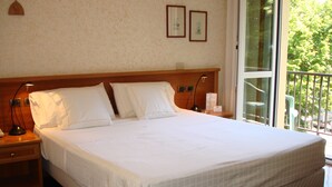 A large bed with white linens, a wooden headboard, a nightstand with a lamp, and a window with a view of greenery. - Ariston (Acqui Terme)