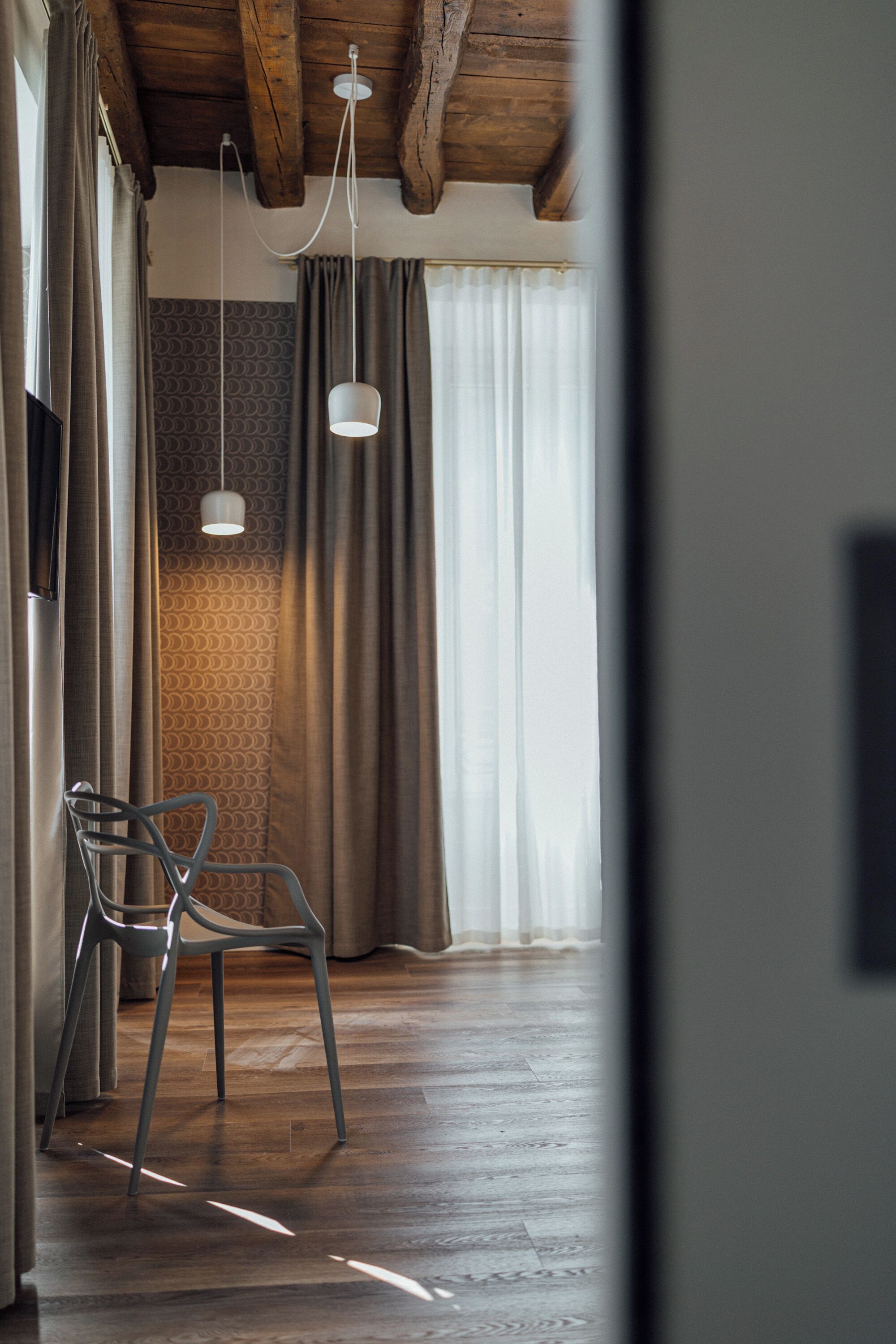 Une pièce avec un plancher en bois, une chaise, un luminaire suspendu et une fenêtre avec des rideaux.