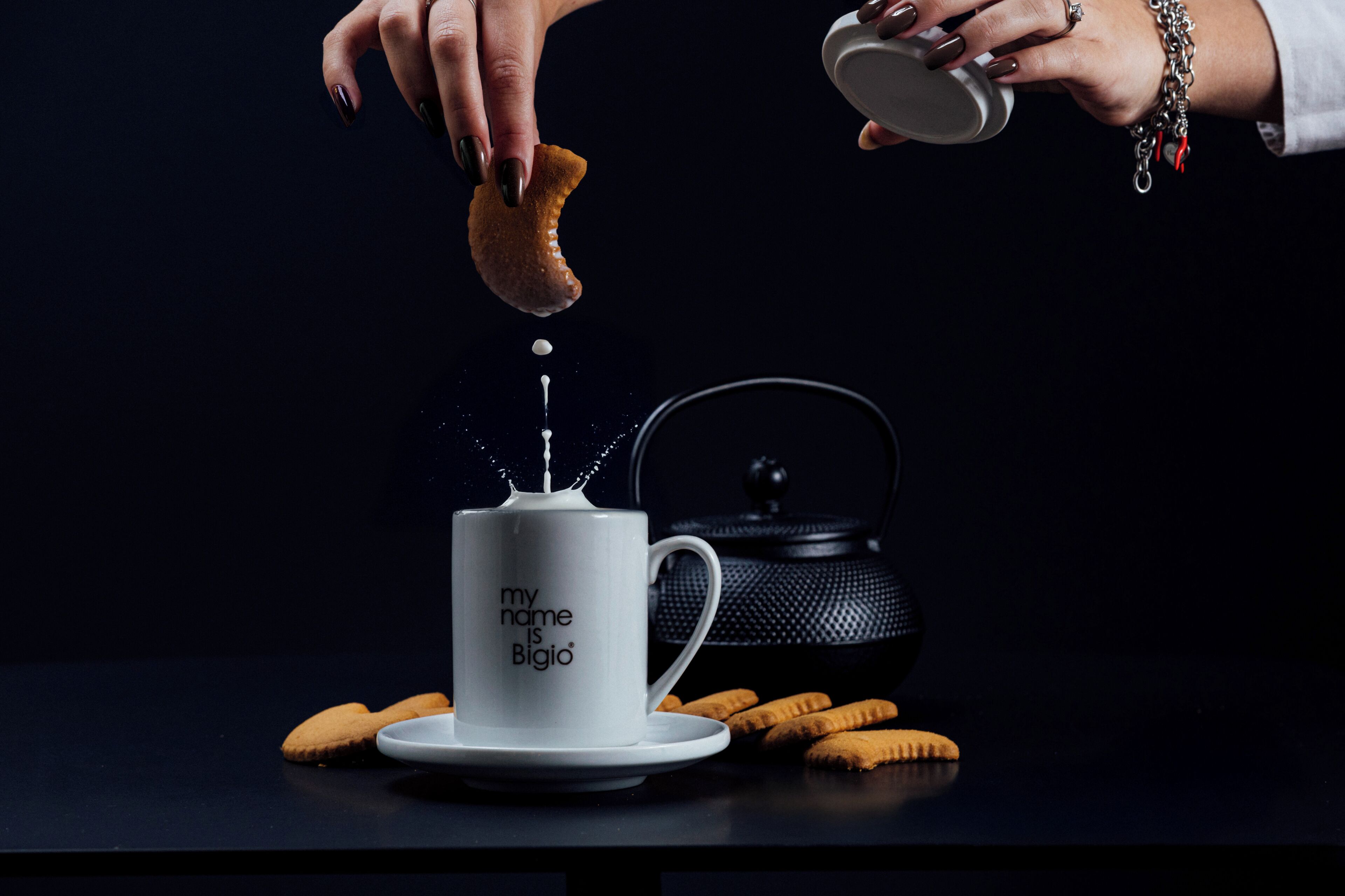 A white mug with text, a teapot, and a biscuit on a dark surface.