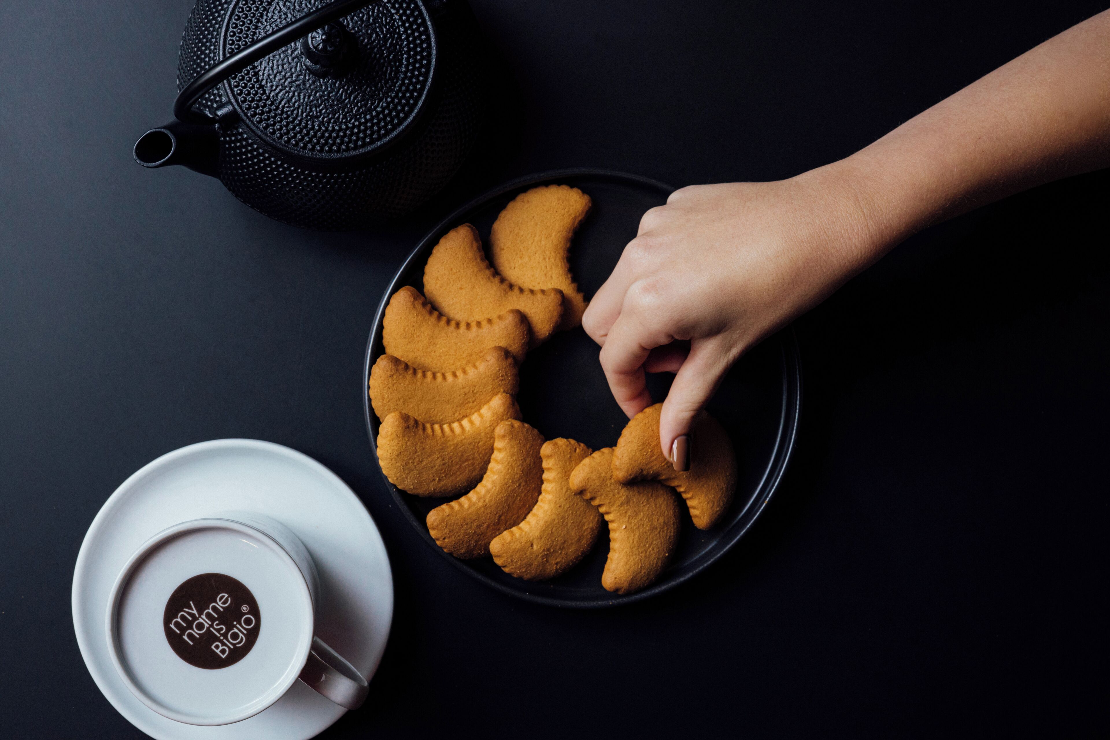 A black teapot, a plate with cookies, and a cup with a logo on a dark surface.