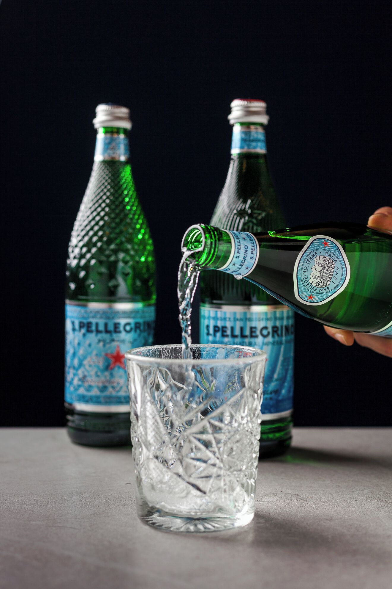 A hand pouring a clear liquid from a green bottle into a clear glass.