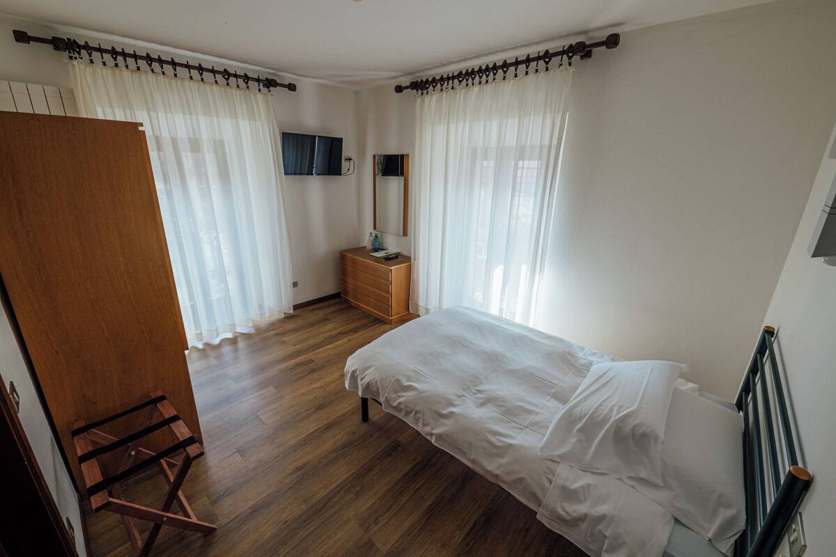 Un dormitorio con suelo de madera, una cama con ropa de cama blanca, un aparador de madera, un espejo y cortinas traslúcidas.