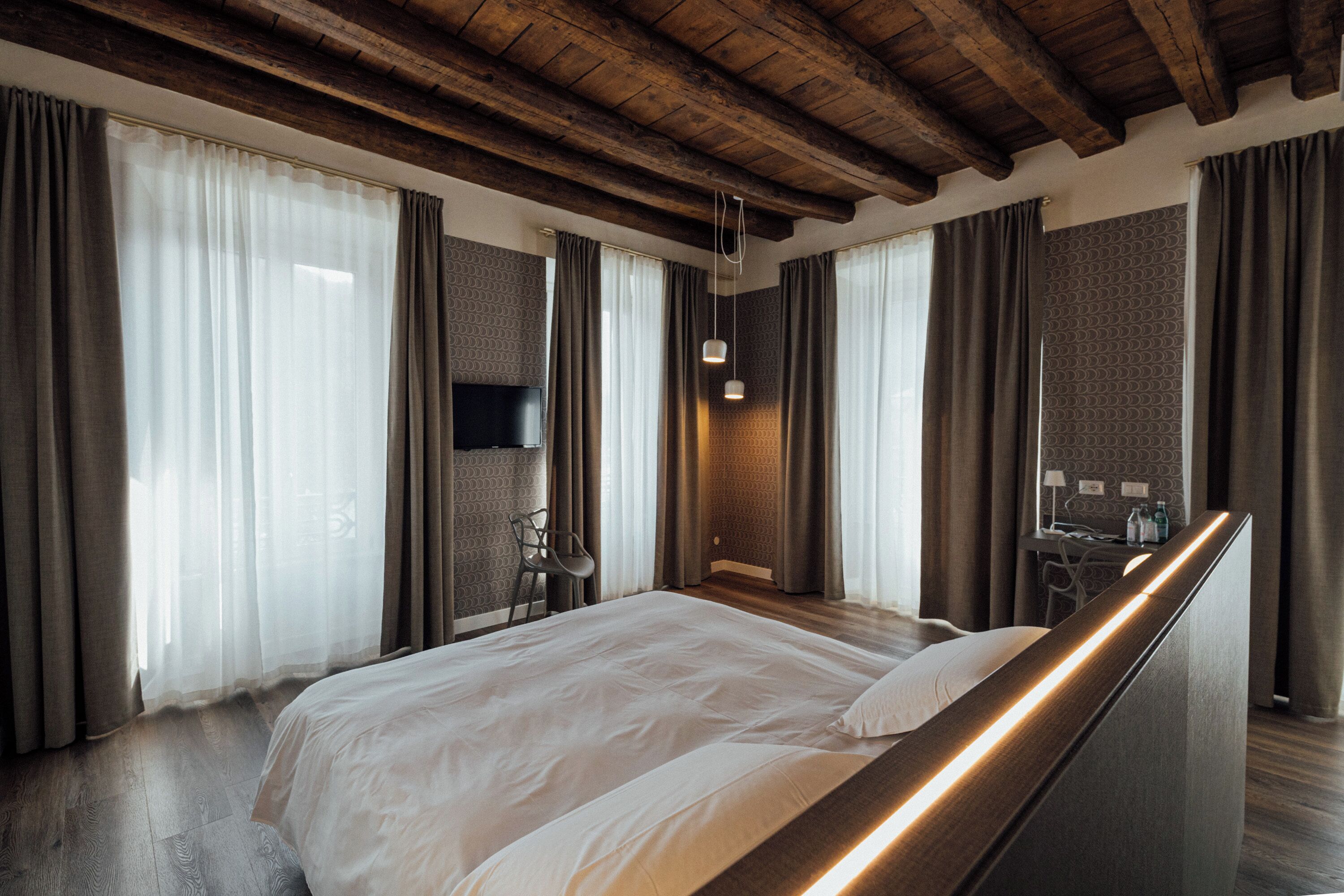 A modern hotel room with a wooden ceiling, a large bed, a desk with a chair, and a TV.