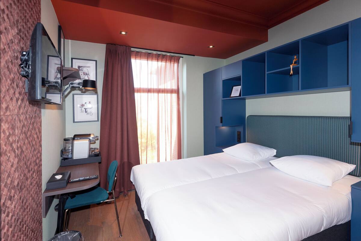 A hotel room with a bed, a desk with a chair, a TV, and a shelf with decorative items.