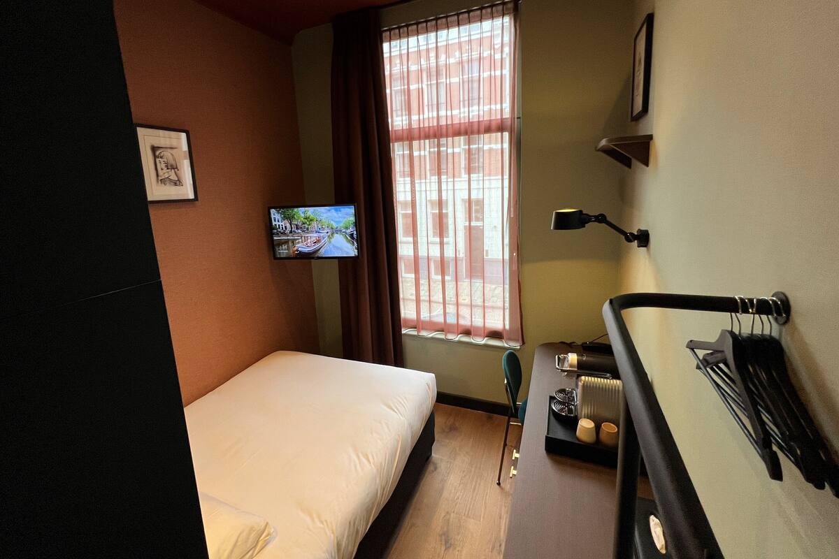 A hotel room with a bed, a TV mounted on the wall, a window with blinds, a desk with a lamp, and a chair.