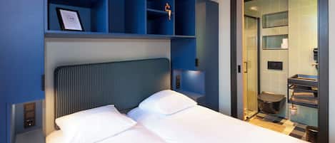 A hotel room with a bed, a headboard, a shelf with a framed picture, a wall-mounted light, and a bathroom visible through an open door.