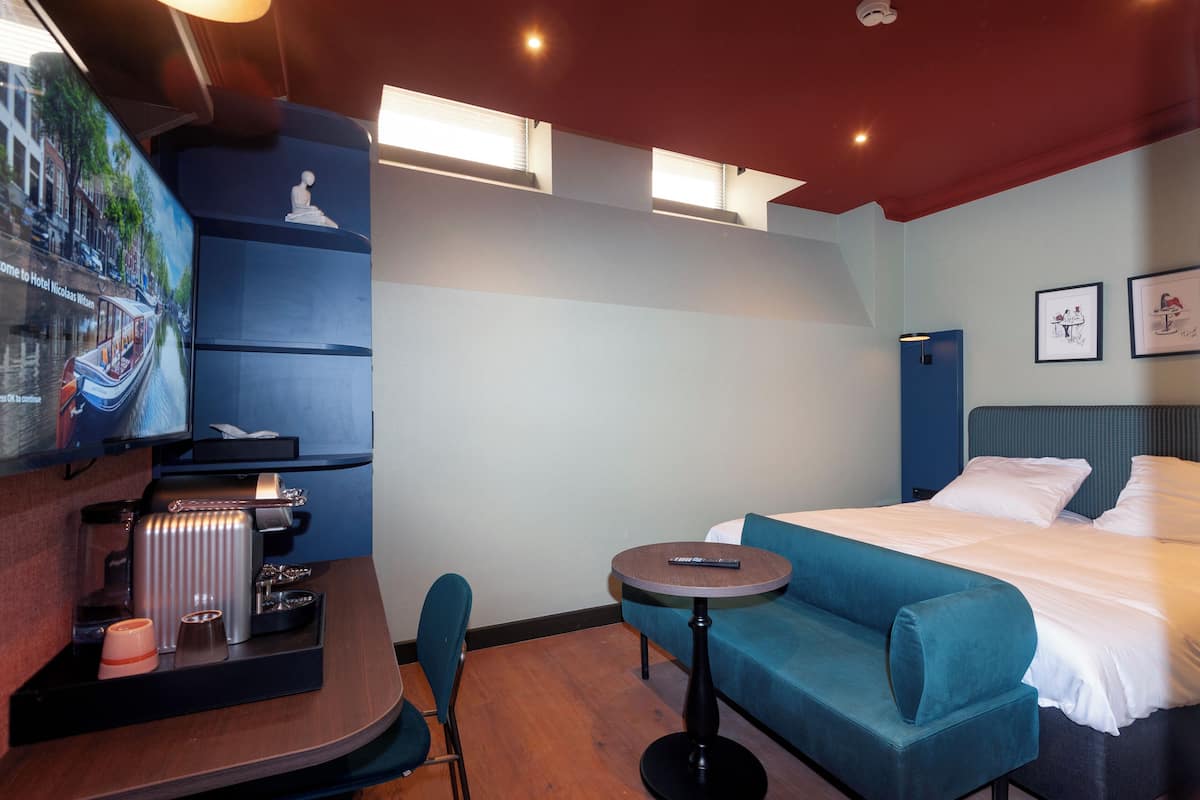 A hotel room with a bed, a sofa, a small table, a chair, a coffee machine, and a TV screen displaying a cityscape.