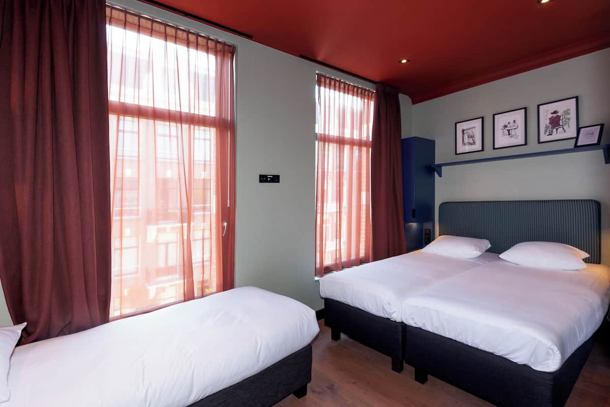A hotel room with two beds, large windows with curtains, a headboard, and wall-mounted artwork.