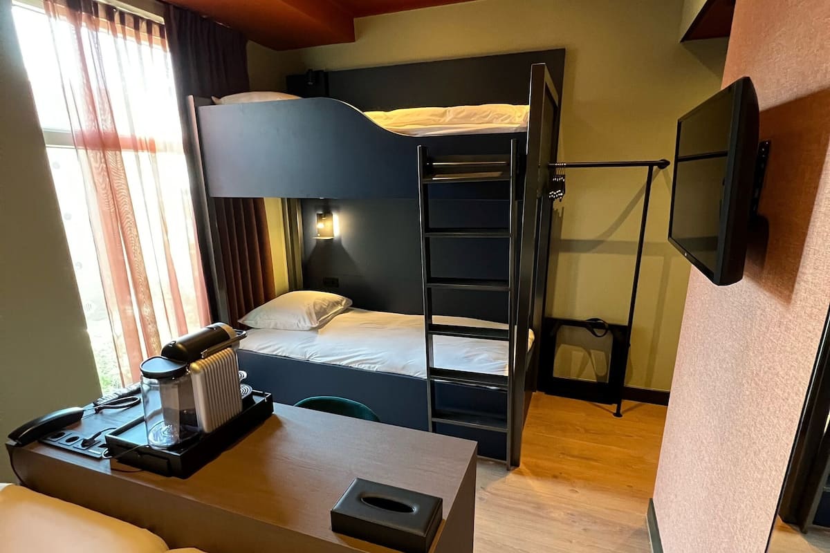 A hotel room with a bunk bed, a desk with a coffee maker, a TV mounted on the wall, and a window with curtains.