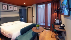 Hotellihuone, jossa on suuri sänky, pieni pöytä, jossa on kasvi, työpöytä tuolilla ja televisio seinällä.