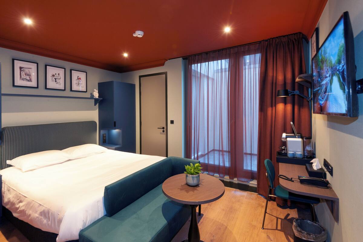 A hotel room with a large bed, a small table with a plant, a desk with a chair, and a TV on the wall.