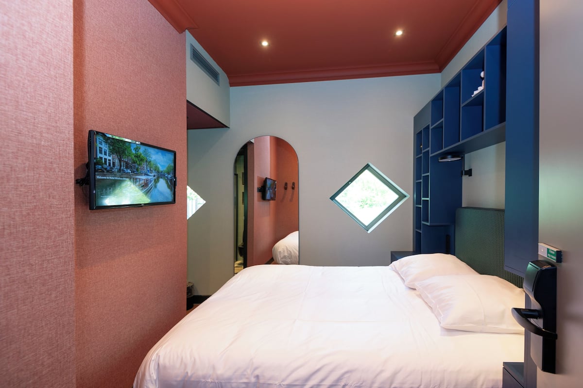 A hotel room with a bed, a TV mounted on the wall, a window, and built-in shelving.