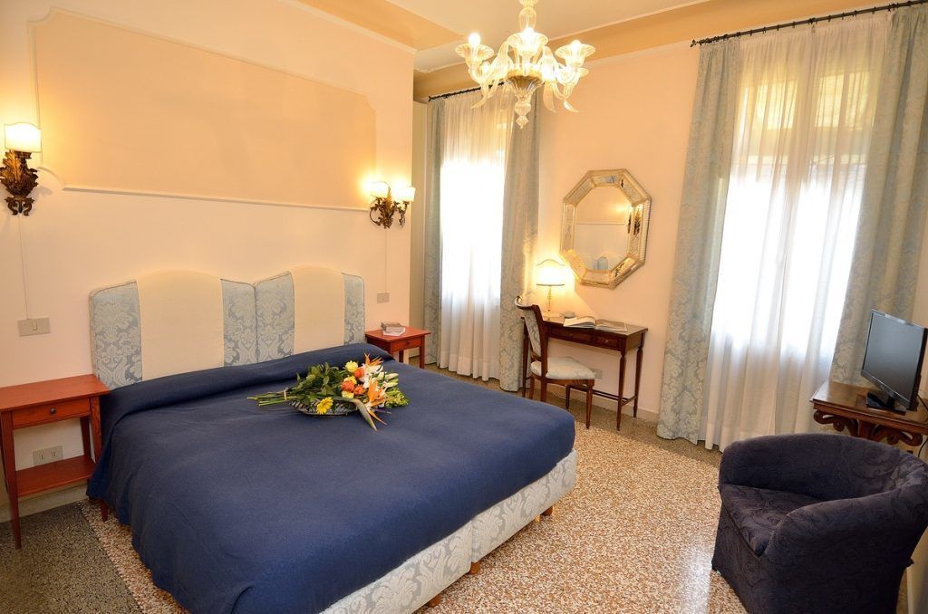 A hotel room with a large bed, a desk, a chair, a television, and a chandelier.
