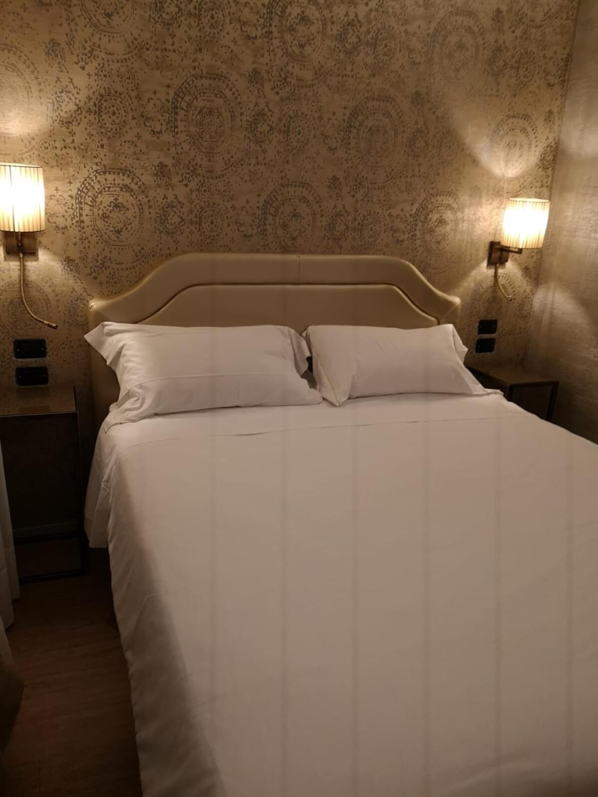 Un lit bien fait, avec du linge de lit blanc et des oreillers, deux lampes de chevet et un papier peint à motifs.
