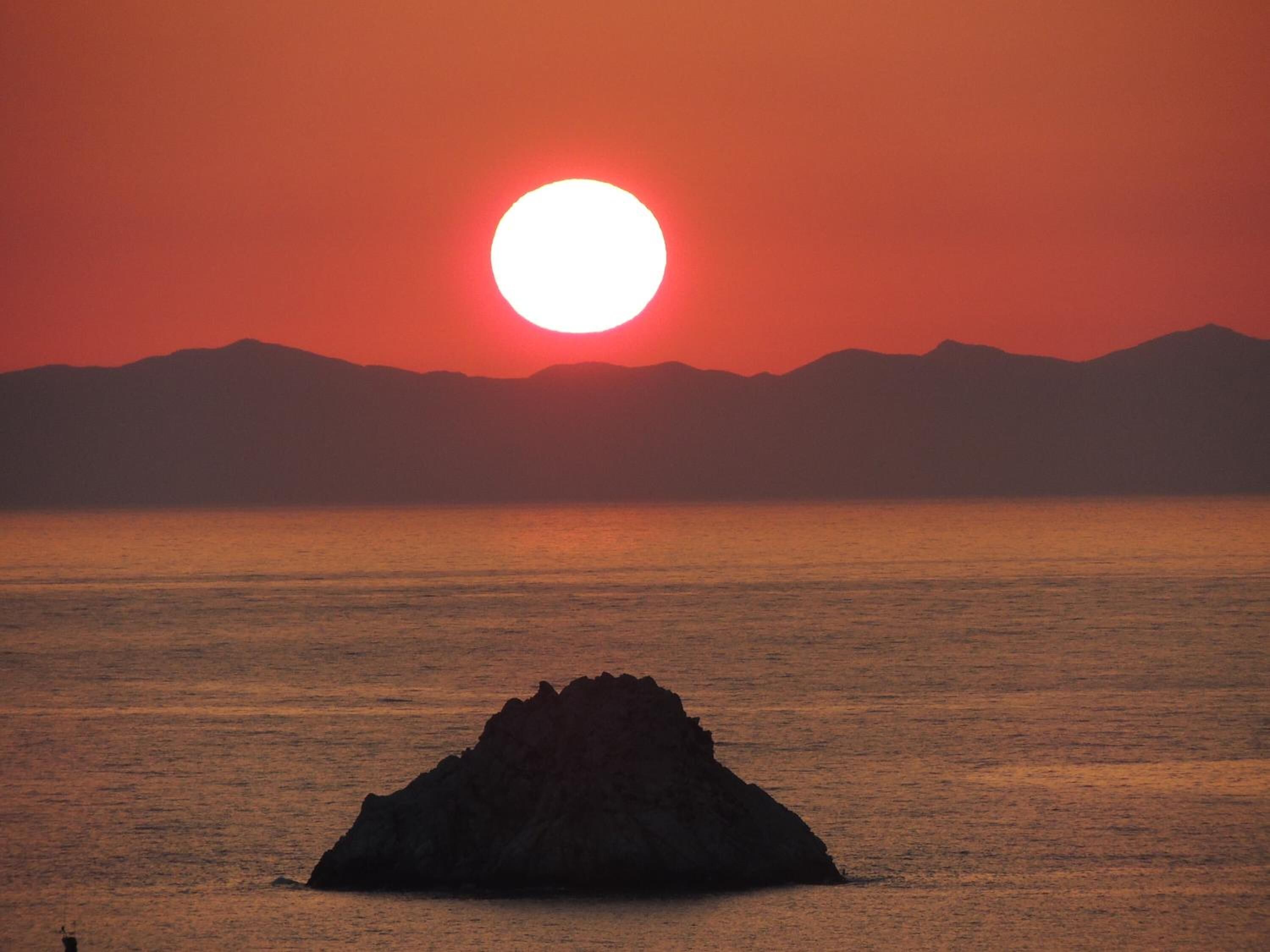 A sunset over a calm sea with a prominent rock formation in the foreground.