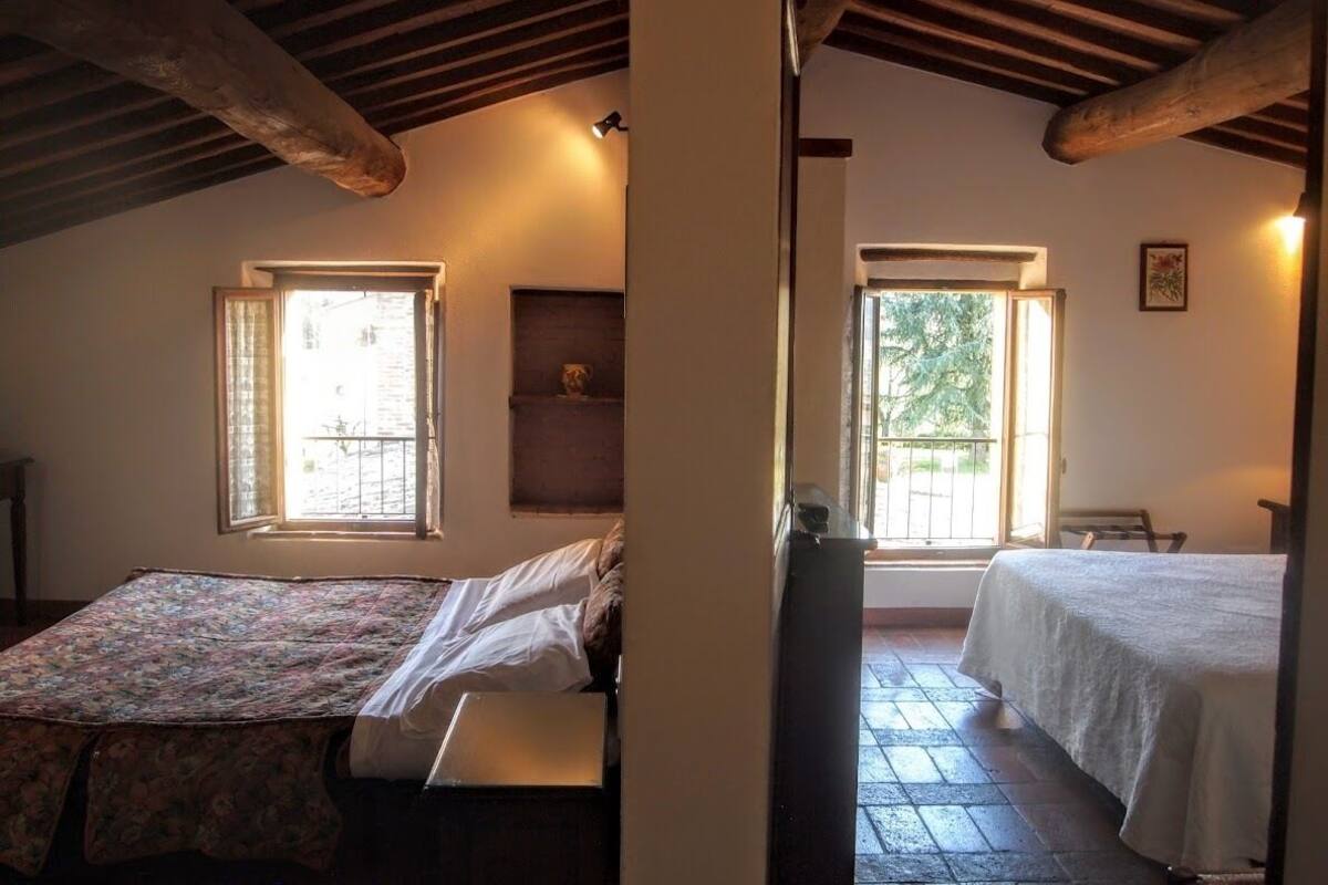 Quarto com duas camas, uma janela, uma prateleira com objeto decorativo e uma porta que dá para outro espaço.