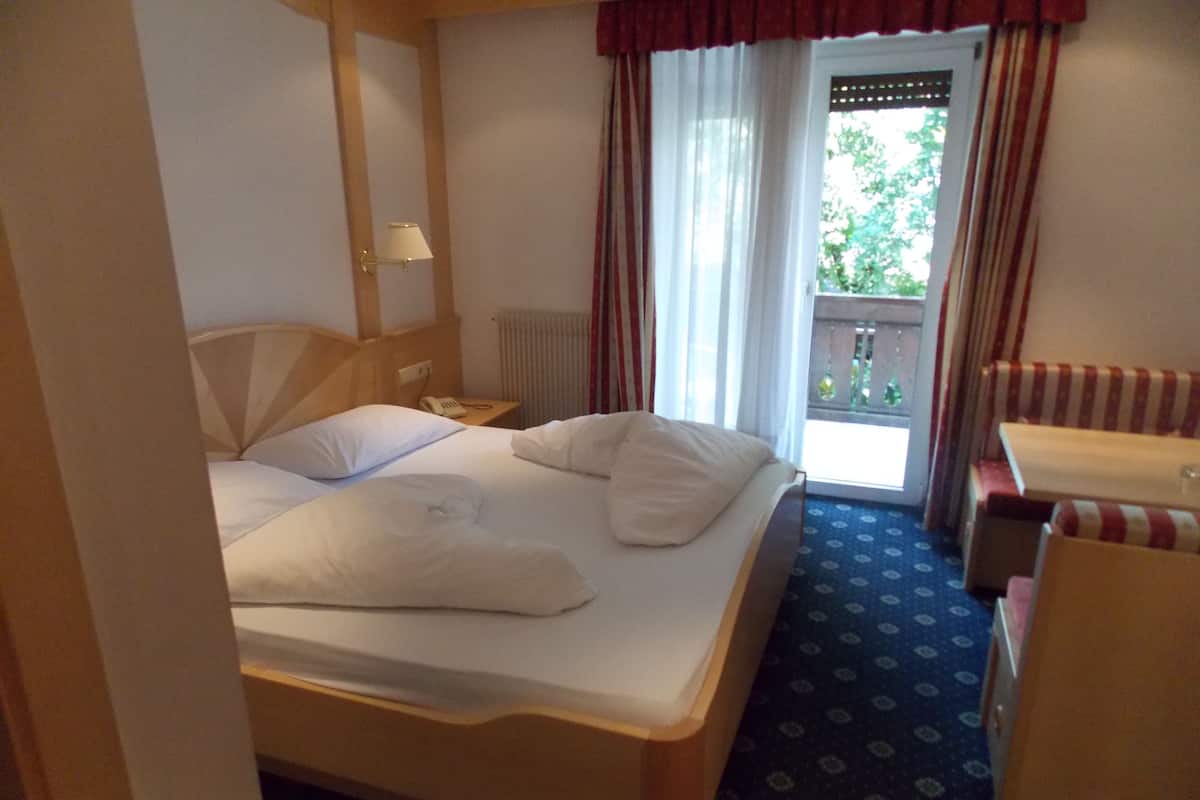 A hotel room with a bed, a bedside table with a lamp, a window with curtains, and a view of the outdoors.
