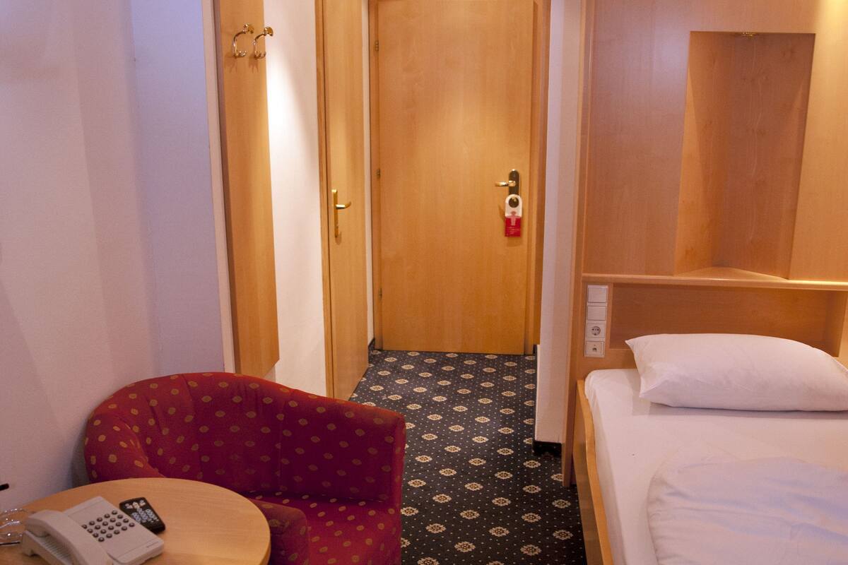 A hotel room with a single bed, a small round table with a telephone, a red armchair, and a patterned carpet.