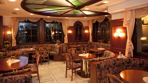Een restaurant met ronde tafels en beklede zitjes, grote ramen voorzien van gordijnen en wandlampen.