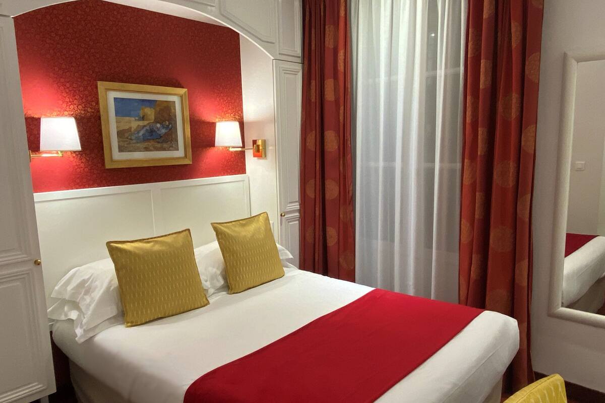 Habitación de hotel con una cama grande, ropa de cama roja y blanca, dos almohadas doradas, un cuadro en la pared y una ventana con cortinas rojas.