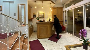 A hotel lobby with a reception desk, wooden cabinets, a red mat, a staircase with a white railing, and a potted orchid.