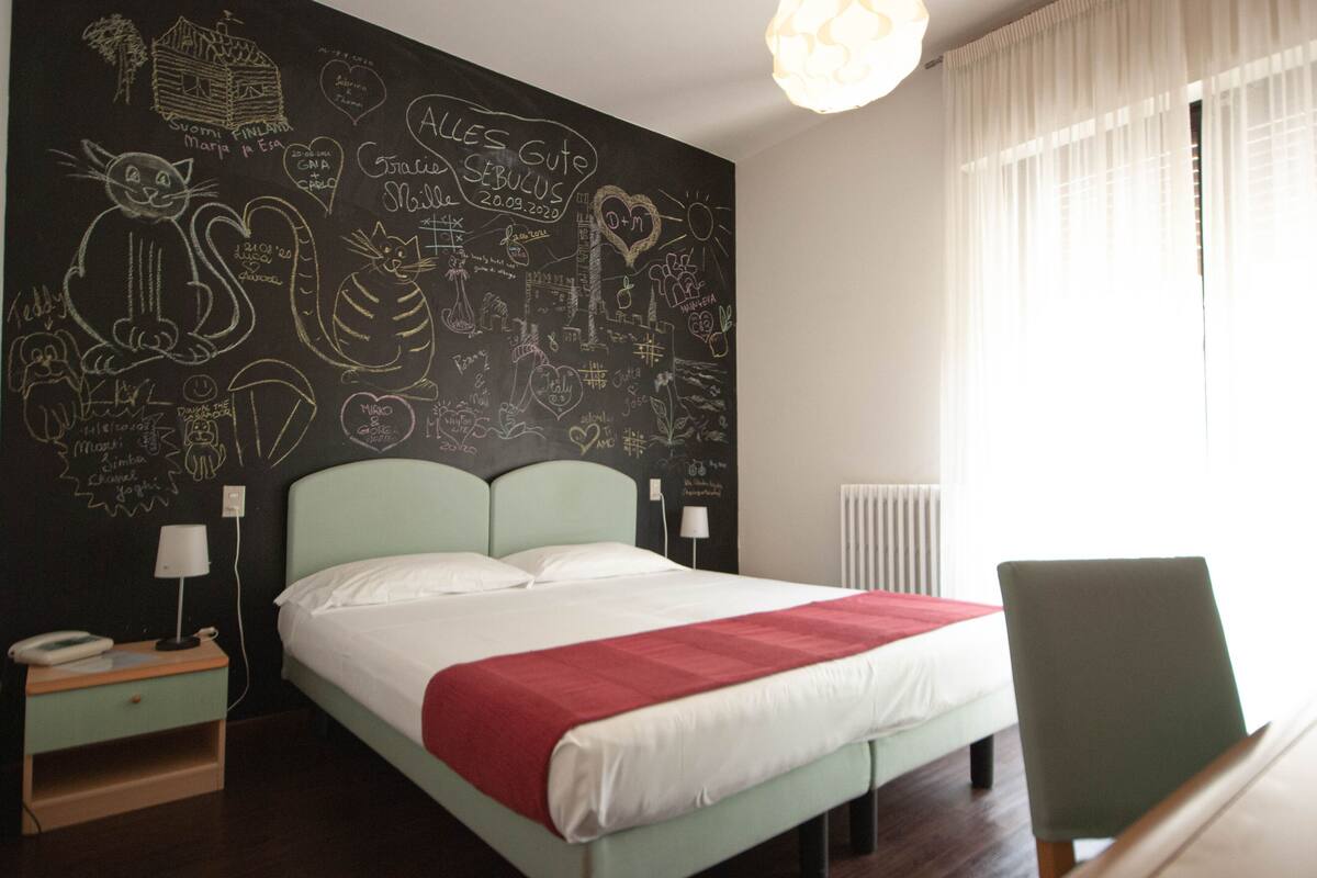 A bedroom with a bed, a nightstand, a chair, and a wall with drawings and writings.