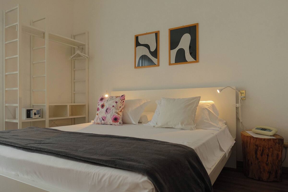 A neatly made bed with a gray blanket, white pillows, and a floral pillow. There is a wooden nightstand with a lamp and a phone. A white wardrobe with a ladder is to the left, and two abstract paintings hang on the wall above the bed.