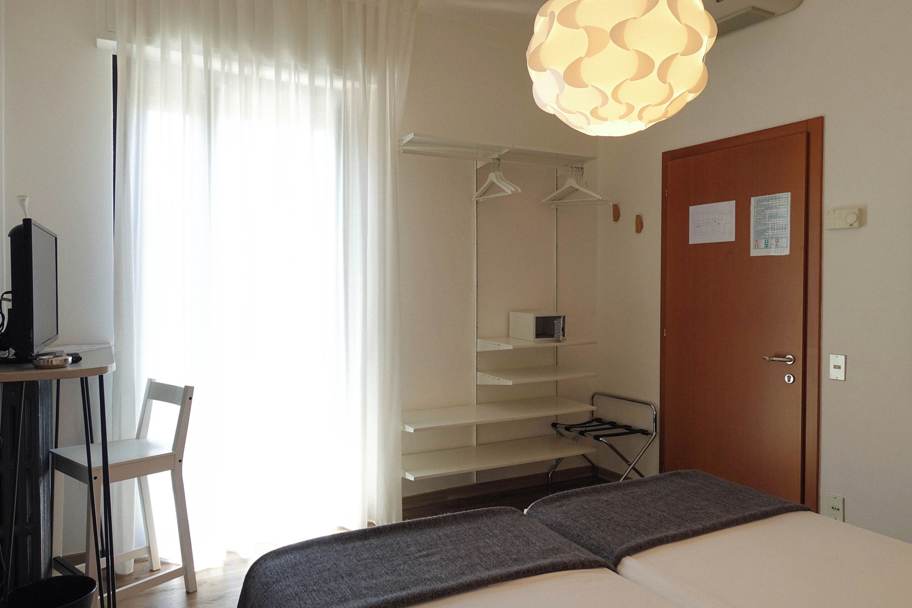Ein Hotelzimmer mit Bett, Schreibtisch mit Stuhl, Kleiderschrank mit Kleiderbügeln, einer Tür mit Durchschlag und einer Hängeleuchte.