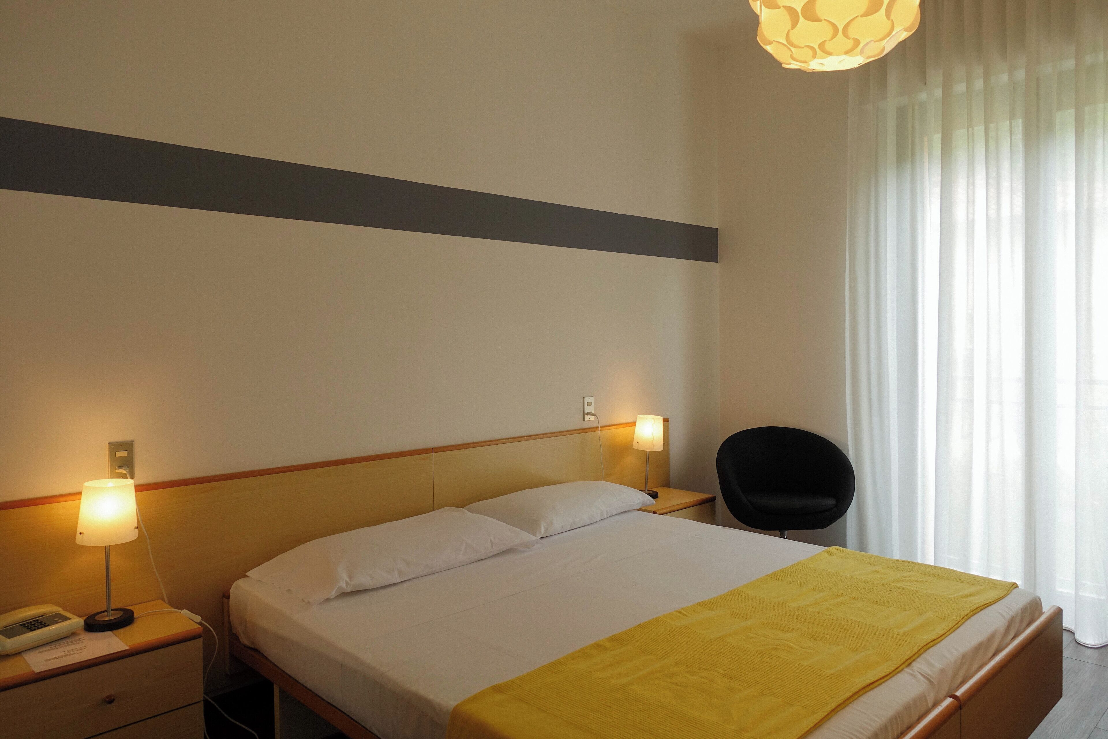 A hotel room with a bed, bedside lamps, a chair, and a window with curtains.