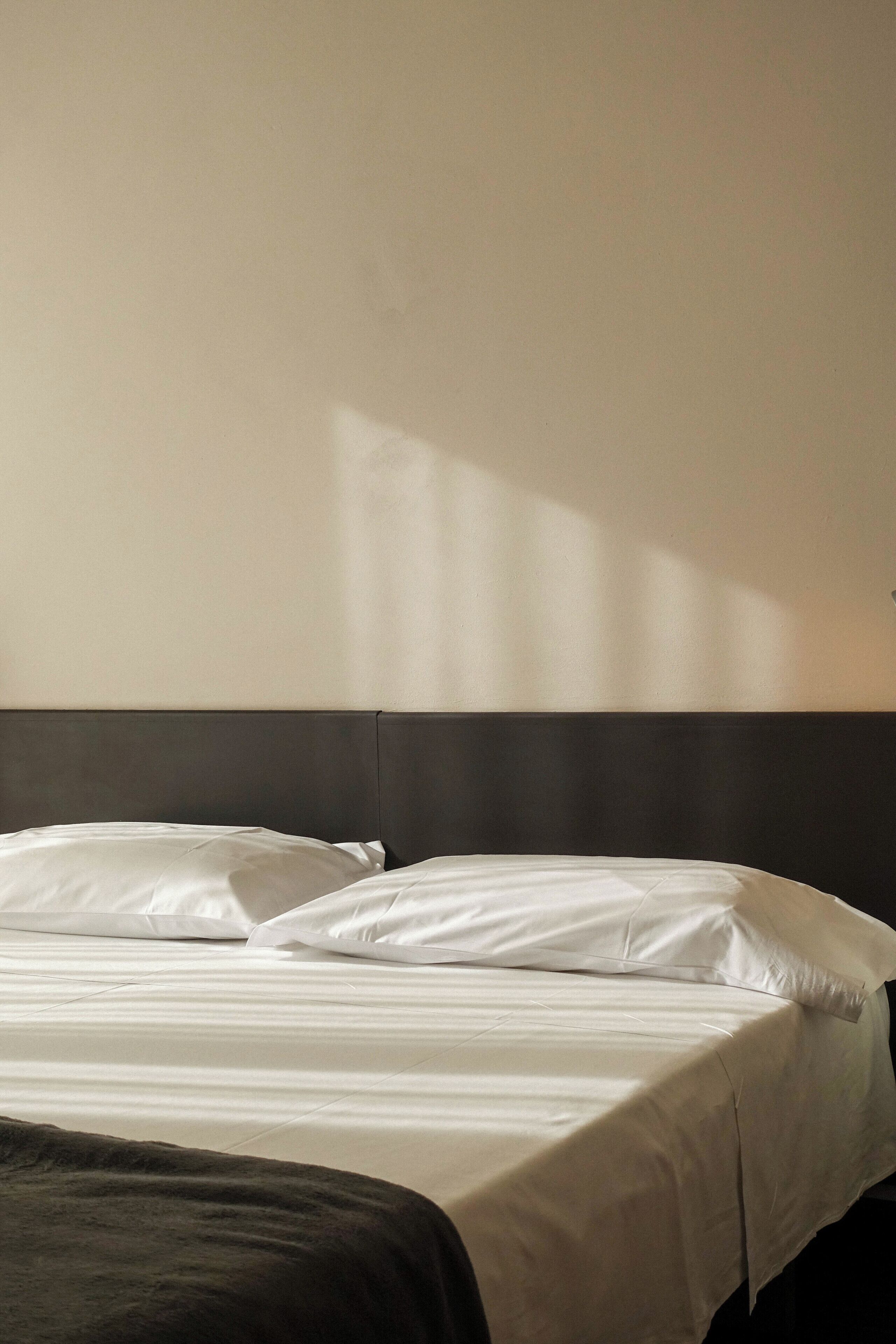 Un lit avec du linge de lit blanc et une tête de lit sombre, dans une pièce dont le mur est d’une couleur neutre.