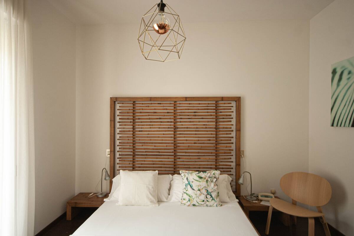A bedroom with a wooden slat headboard, a bed with white linens, a chair, and a geometric pendant light.