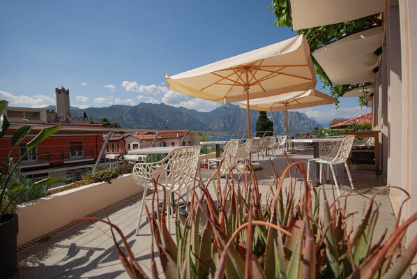 An outdoor terrace with white chairs and a large beige umbrella, overlooking a mountainous landscape. - Hotel Peler (Malcesine)