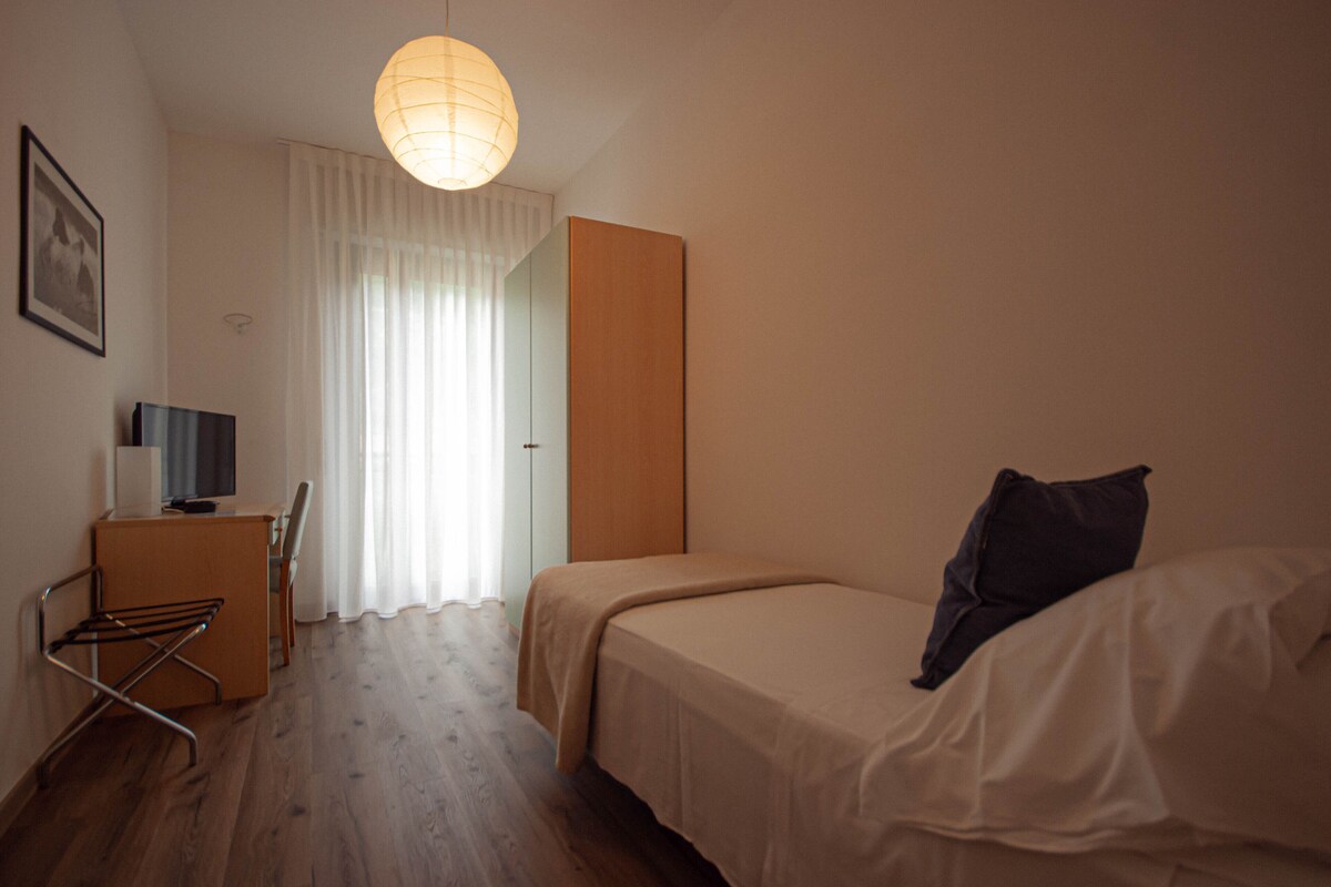 A small, well-lit room with a bed, a desk, and a chair.