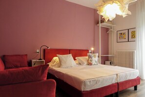A bedroom with a red bed, white bedding, a red chair, a bedside table with a lamp, and a radiator. - Hotel Peler (Malcesine)