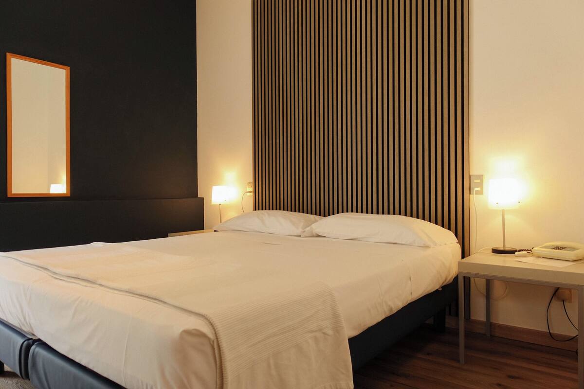 A neatly made bed with white linens, a bedside table with a lamp, and a wall-mounted mirror.