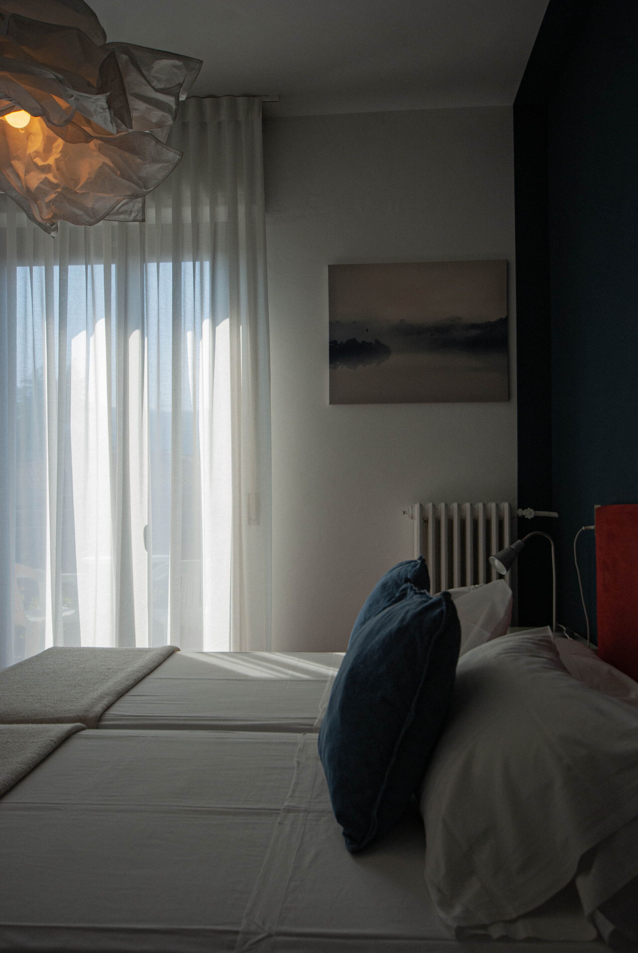 A bedroom with a bed, a radiator, a window with sheer curtains, and a painting on the wall.